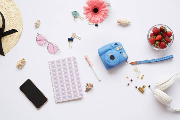 Above view of womens stuff on white background: instant camera, smartphone, notepad, pink sunglasses, straw hat and pins on table - Foto, Bild