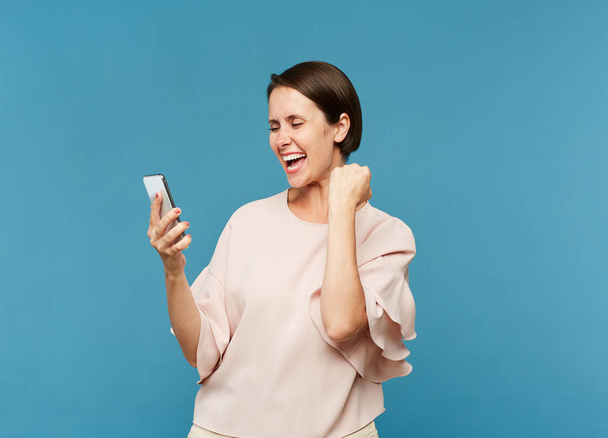 Young ecstatic female looking at smartphone screen while expressing gladness in isolation - 写真・画像