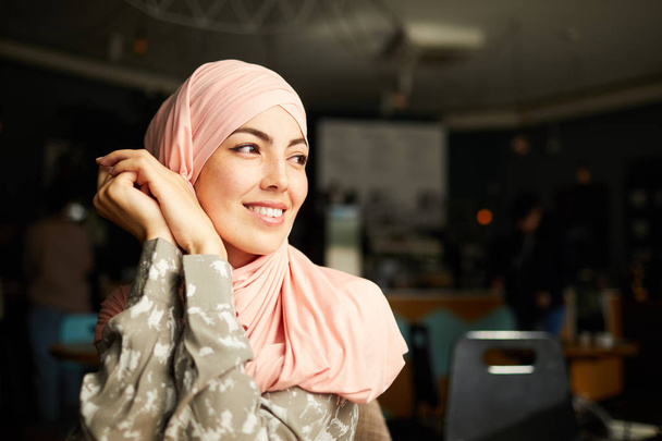 Całkiem uśmiechnięta młoda muzułmańska kobieta siedzi przy stole w kawiarni i przegląda okno - Zdjęcie, obraz