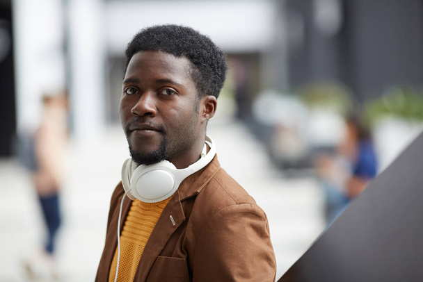 Πορτραίτο του σοβαρού όμορφου νεαρού μαύρου άντρα με ενσύρματα ακουστικά στο λαιμό που στέκονται στο λόμπι - Φωτογραφία, εικόνα
