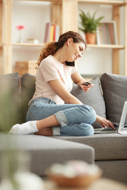 Uśmiechnięta młoda kobieta z kręcone włosy siedzi na kanapie i wstawianie numer karty kredytowej do zakupu podczas rozmowy na telefon komórkowy - Zdjęcie, obraz
