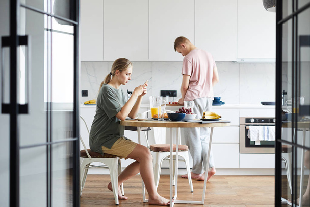 Young wife texting in smartphone by brunch while her husband cooking in the kitchen - Photo, Image