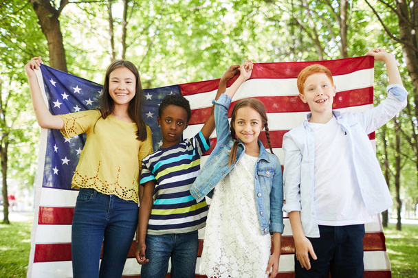 Группа позитивных мультиэтнических детей в повседневных нарядах, стоящих в лесу и держащих американский флаг вместе во время празднования Дня независимости
 - Фото, изображение
