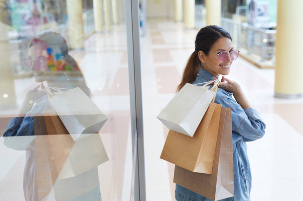 junges lächelndes Mädchen mit pinkfarbener Sonnenbrille spaziert nach erfolgreichem Einkauf mit verschiedenen Taschen durch Einkaufszentrum - Foto, Bild
