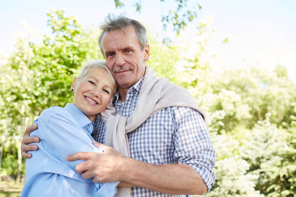 Waist up portrait of loving senior couple embracing and looking at camera while enjoying walk in forest, copy space - Photo, Image