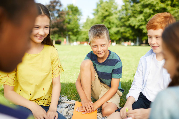 公園の芝生の上でサークルに座って面白いゲームをしている5人の笑顔の小学生 - 写真・画像