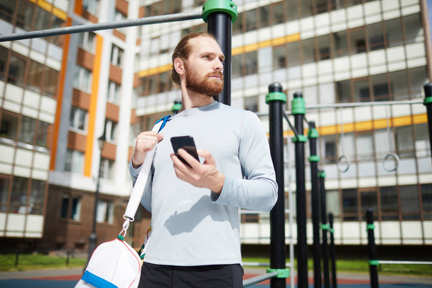Content determined young sportsman with red beard standing at workout area and using modern smartphone while checking exercise task - Photo, Image