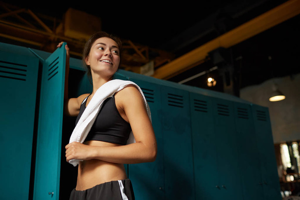 Portrait sombre à angle bas d'une jeune femme souriante debout près d'un casier dans un club de sport et regardant ailleurs, copiant l'espace
 - Photo, image