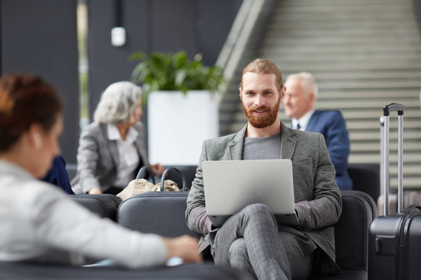 Porträt eines lächelnden, rotbärtigen Unternehmers in grauer Jacke, der auf dem Sofa sitzt und Laptop am Flughafen benutzt - Foto, Bild