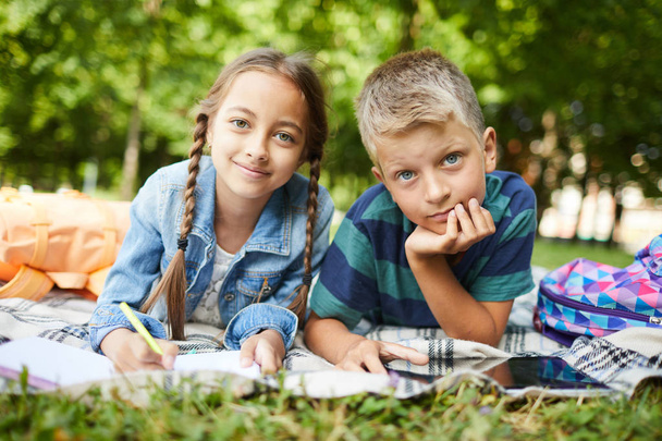 Двоє усміхнених дітей охолоджують після школи на траві в парку, роблячи домашнє завдання разом надворі
 - Фото, зображення