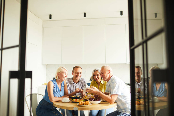 Groep van vrolijke senior mensen zitten rond tafel in de keuken geschoten door open deuren, kopieer ruimte - Foto, afbeelding