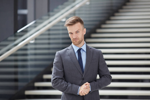 Σοβαρός όμορφος νέος επιχειρηματίας με γκρι κοστούμι στέκεται εναντίον σκάλες στο λόμπι και κοιτάζοντας την κάμερα με σιγουριά - Φωτογραφία, εικόνα