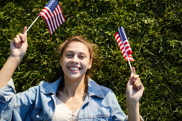 Vue en angle élevé d'une étudiante joyeuse avec un large sourire allongé sur l'herbe et agitant des drapeaux de bâton américains tout en se relaxant le 4 juillet
 - Photo, image