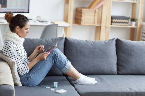 Άρρωστη νεαρή γυναίκα με ουρά πόνι φορώντας ζεστό κασκόλ γύρω από το λαιμό κάθεται στον καναπέ με δοχεία χαπιών και περιήγηση ιατρικές ιστοσελίδες στο tablet στο σπίτι - Φωτογραφία, εικόνα