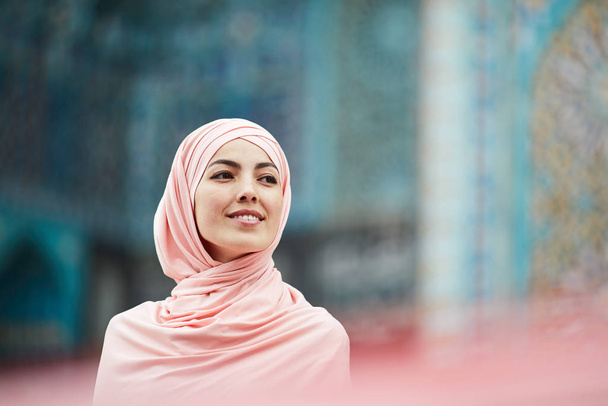 Χαμογελαστή νεαρή αραβική κυρία σε μετάξι χιτζάμπ στέκεται ενάντια διακοσμητική πρόσοψη του κτιρίου και κοιτάζοντας μακριά - Φωτογραφία, εικόνα