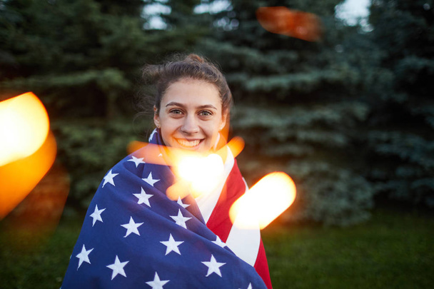 Portrait of happy patriotic American girl wrapped in national flag standing in forest, sparkles effect - Photo, Image