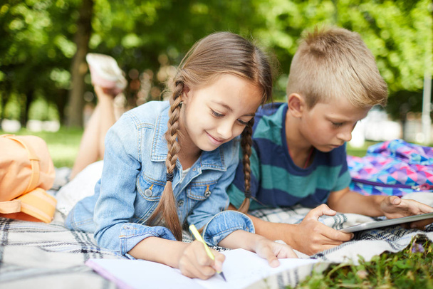 Δύο σχολικά παιδιά ξοδεύουν ελεύθερο χρόνο στο πάρκο κατά τη διάρκεια των καλοκαιρινών διακοπών σχεδιάζοντας και παίζοντας με το tablet - Φωτογραφία, εικόνα