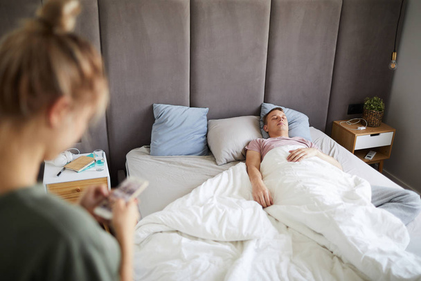 Young tired man sleeping in bed while wife or girlfriend scrolling in his smartphone near by - Photo, image