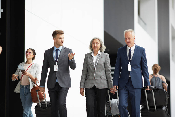 Група багатоетнічних бізнесменів у костюмах, що рухаються вздовж аеропорту з валізами та слухають молодого колегу
 - Фото, зображення