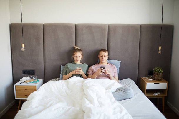 Młody Zdejmowany para leżący w łóżko podczas przy pomocy ich Smartphones za spać - Zdjęcie, obraz