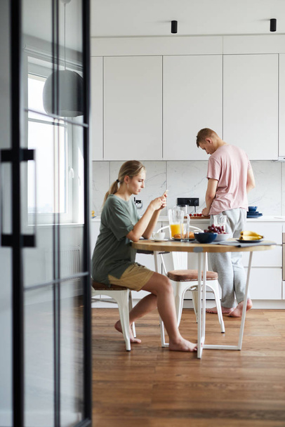 Nuori nainen istuu pöydän ääressä keittiössä, syö aamiaista ja käyttää älypuhelinta, kun hänen miehensä ruoanlaitto
 - Valokuva, kuva