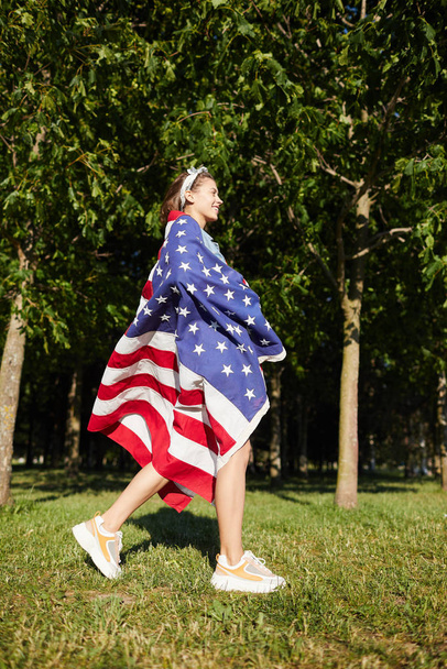 lächelnd aufgeregtes hübsches Mädchen mit Kopftuch und stylischen Turnschuhen, die in eine große amerikanische Flagge gehüllt im Sommerpark spazieren gehen - Foto, Bild