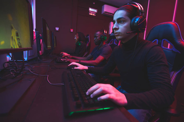 Serious focused multi-ethnic e-sports gamers in dark clothes sitting at table and playing online game via network - Foto, Imagen