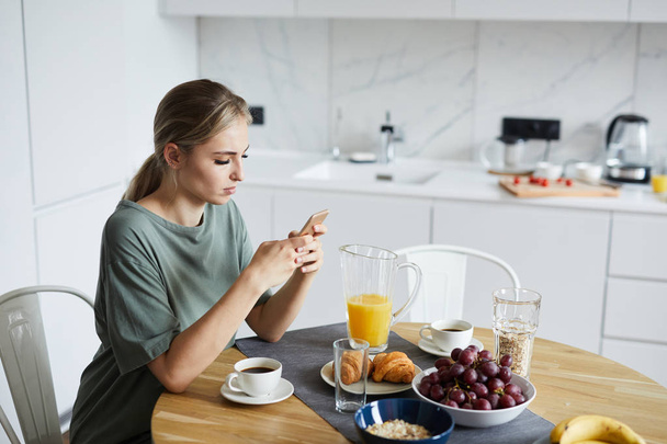 Młoda atrakcyjna kobieta przy użyciu smartfona, podczas gdy śniadanie lub brunch w kuchni - Zdjęcie, obraz