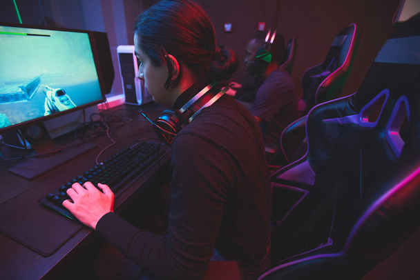 Konzentrierter Jude mit Headset am Hals sitzt am Tisch im dunklen Raum und spielt Computernetzwerk - Foto, Bild