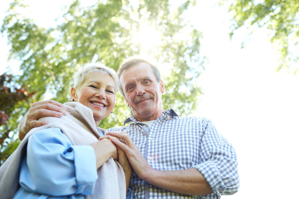 Low angle portrait of happy senior couple looking down at camera and smiling while walking in Summer park, copy space - Foto, Imagen