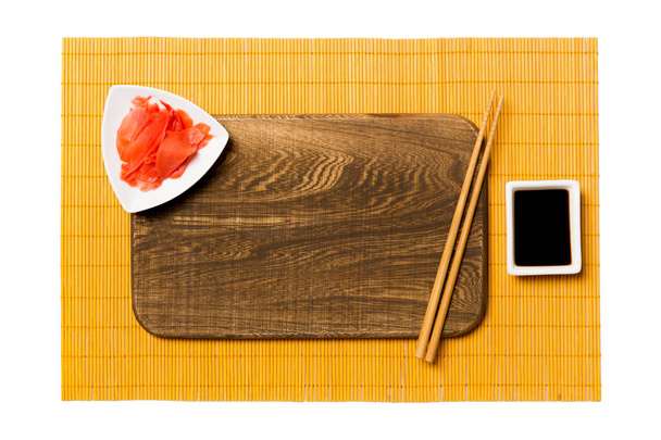 Prázdná obdélníková hnědá Dřevěná deska s hůlky pro sushi, zázvorové a sójové omáčky na žlutém podkladu. Pohled shora s místem kopírování pro návrh - Fotografie, Obrázek