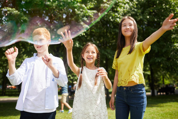 Cheerful carefree friends in casual clothing standing in city park and gesturing hands while looking at soap bubbles - Photo, Image