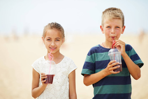 Content little friends in casual outfits standing on beach and drinking fruit juice from straws while looking at camera - Fotografie, Obrázek