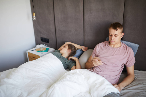 Young suspicious man reading messages of his wife in her smartphone while sitting in bed - Foto, immagini