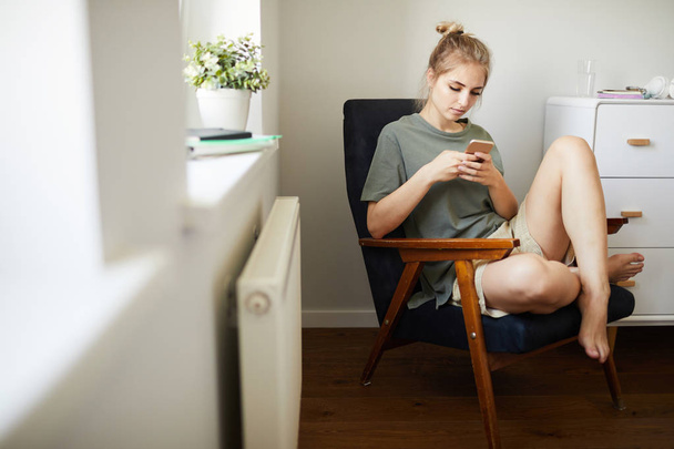 Симпатична дівчина в повсякденному одязі дивиться відео в смартфоні, розслабляючись в кріслі в спальні
 - Фото, зображення