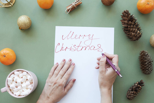 Γυναικεία χέρια που υπογράφουν Χριστουγεννιάτικη κάρτα με στυλό στο πράσινο τραπέζι με χριστουγεννιάτικα στολίδια και λιχουδιές - Φωτογραφία, εικόνα