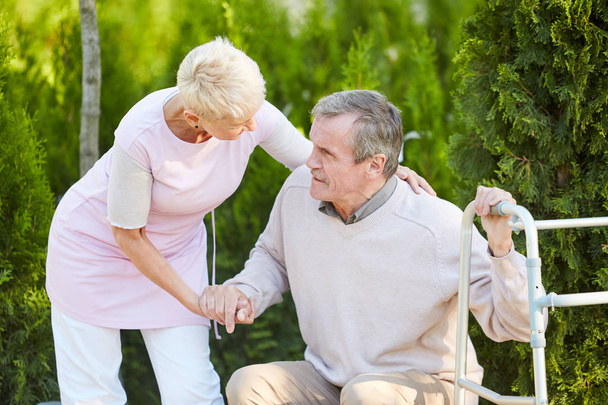 Portrait of caring nurse helping senior man get up from park bench in rehabilitation center, copy space - Photo, Image