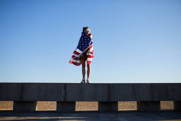 Smiling attractive young woman in sunglasses wrapped in American flag standing on concrete plate and contemplating landscape - Photo, Image
