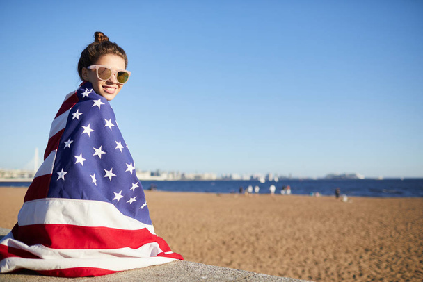 Retrato de mulher jovem atraente feliz com cabelo bun usando óculos de sol cobertos de grande bandeira americana sentado em laje de concreto enquanto passa tempo na praia
 - Foto, Imagem