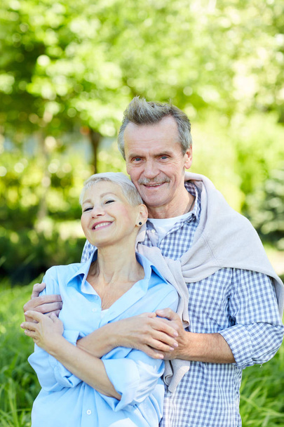Μέση επάνω πορτρέτο του ευτυχισμένο γηραιότερος ζευγάρι Αγκαλιάζοντας και κοιτάζοντας την κάμερα, απολαμβάνοντας το περπάτημα στο καλοκαιρινό πάρκο - Φωτογραφία, εικόνα