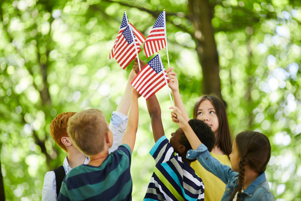 Grupo de niños patrióticos multiétnicos de pie en círculo y levantándonos banderas pegadas juntos
 - Foto, Imagen