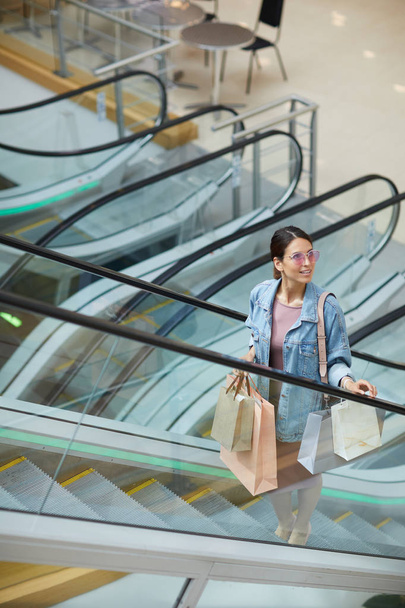 Sonriente joven de moda en gafas de sol de color rosa de pie en la escalera mecánica en el centro comercial y la celebración de bolsas de compras
 - Foto, Imagen