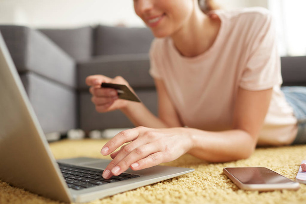 Close-up van lachende jonge vrouw liggend op tapijt en het gebruik van creditcard en laptop terwijl het doen van online winkelen thuis - Foto, afbeelding