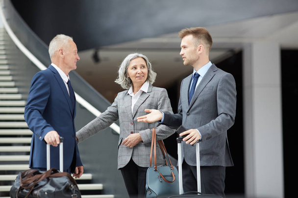 Группа бизнес-профессионалов в формальной одежде, стоящих в аэропорту и держащих чемоданы во время разговора
 - Фото, изображение