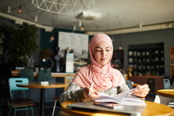 Serious young Muslim woman sitting at cafe table, reading book and taking notes - 写真・画像