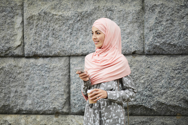 Pozytywny młody środkowy wschód Pani w różowy lato hidżab chodzenie przez ulica i picie Takeout Kawa podczas przy pomocy gadżet u przesuwać - Zdjęcie, obraz