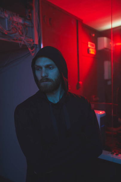 Serious handsome young bearded hacker in hoodie leaving server room with broken Ethernet hub and red light in background - Foto, imagen