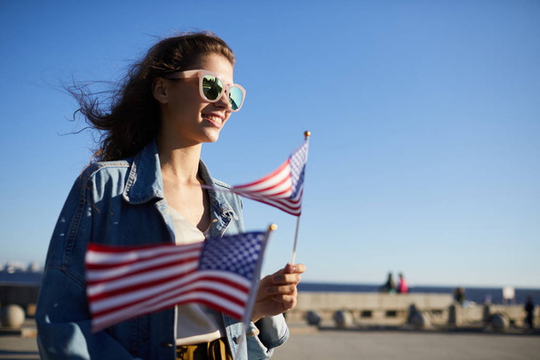 Radostně atraktivní městská dívka v slunečních brýlích na přehlídce na den nezávislosti a drží nám vlajkové vlajky - Fotografie, Obrázek