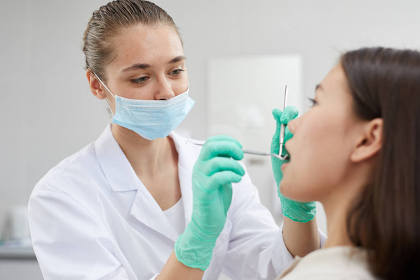 Portrait of young female dentist wearing protective mask while examining patient in clinic, copy space - Фото, изображение