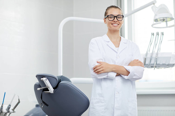 Портрет молодой женщины-врача в очках, улыбающейся в камеру во время позирования с креслом-стоматологом, пространство для копирования
 - Фото, изображение